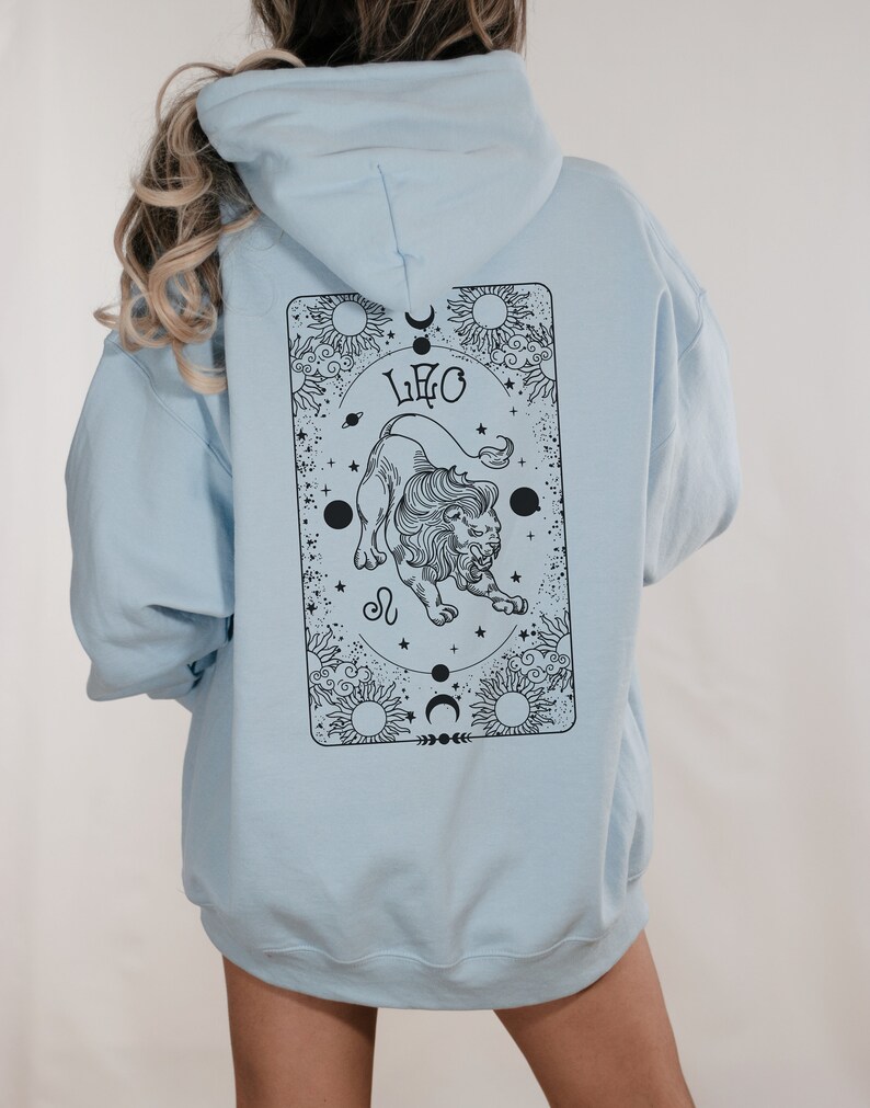 May include: Light blue hoodie with a black Leo zodiac design on the back. The design features a lion, the word "LEO", suns, moons, and stars. The hoodie has a hood and long sleeves.