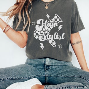 May include: A dark gray t-shirt with a white graphic design of a checkered racing flag with the text "Hair Stylist" in a retro font. The graphic also includes lightning bolts and stars.