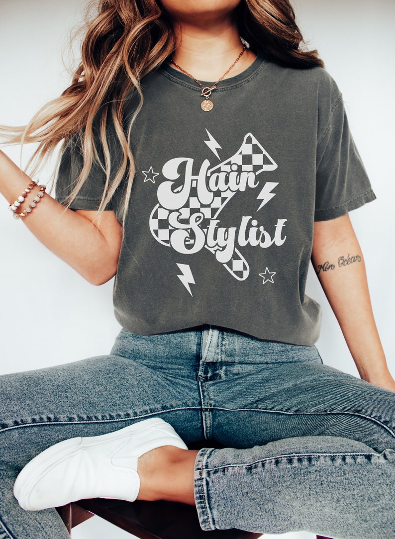 May include: A dark gray t-shirt with a white graphic design of a checkered racing flag with the text "Hair Stylist" in a retro font. The graphic also includes lightning bolts and stars.