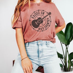 May include: A dusty rose-colored t-shirt featuring a circular graphic with a guitar, stars, and the text "RAISED ON 90'S COUNTRY" in a distressed style. The shirt is paired with light-wash blue jeans. The overall aesthetic is casual and country-themed.
