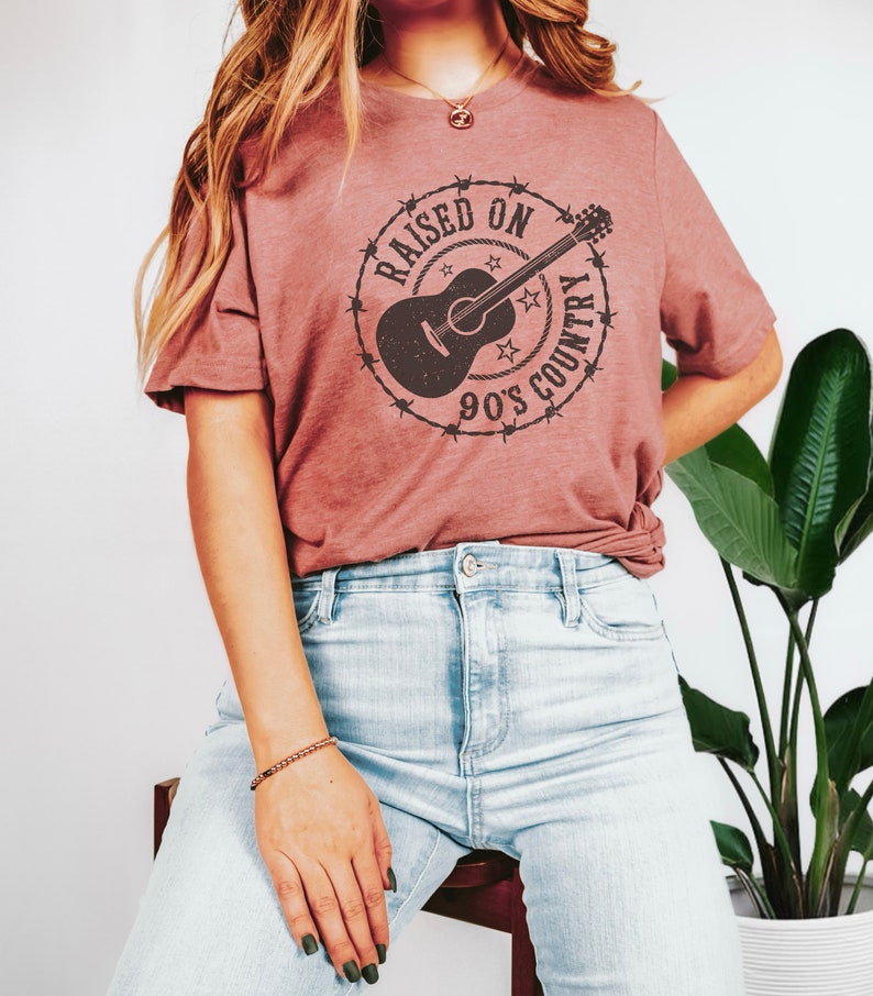 May include: A dusty rose-colored t-shirt featuring a circular graphic with a guitar, stars, and the text "RAISED ON 90'S COUNTRY" in a distressed style. The shirt is paired with light-wash blue jeans. The overall aesthetic is casual and country-themed.