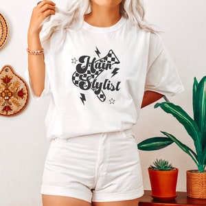 May include: White t-shirt with a black and white checkered graphic that says "Hair Stylist" with lightning bolts and stars.