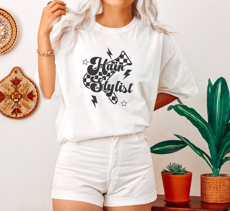 May include: White t-shirt with a black and white checkered graphic that says "Hair Stylist" with lightning bolts and stars.