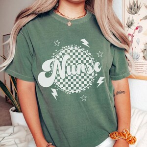 K&ouml;nnte beinhalten: Ein gr&uuml;nes T-Shirt mit einem wei&szlig;en karierten Kreis und dem Wort "Nurse" in einer Retro-Schrift. Der Kreis ist von wei&szlig;en Sternen und Blitzen umgeben.