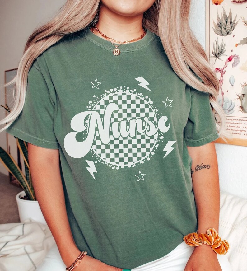 K&ouml;nnte beinhalten: Ein gr&uuml;nes T-Shirt mit einem wei&szlig;en karierten Kreis und dem Wort "Nurse" in einer Retro-Schrift. Der Kreis ist von wei&szlig;en Sternen und Blitzen umgeben.
