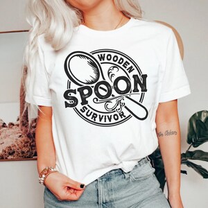May include: White t-shirt with a black and white graphic design of a wooden spoon with the text "Wooden Spoon Survivor".