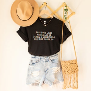 May include: Black t-shirt with the text "THIS BODY CAME WITH A LOT OF TERMS & CONDITIONS I DID NOT AGREE TO" in rainbow colors. The shirt is paired with denim shorts and a straw bag.
