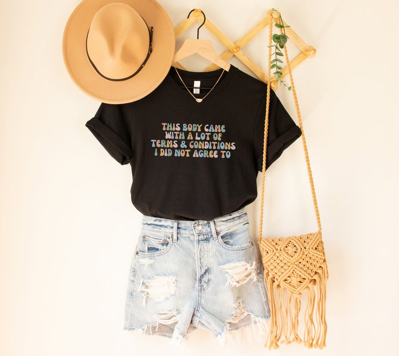 May include: Black t-shirt with the text "THIS BODY CAME WITH A LOT OF TERMS & CONDITIONS I DID NOT AGREE TO" in rainbow colors. The shirt is paired with denim shorts and a straw bag.