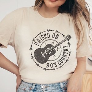 May include: Cream-colored t-shirt featuring a distressed graphic. The design includes a guitar encircled by barbed wire, stars, and the text "RAISED ON 90'S COUNTRY". A casual, short-sleeved tee.