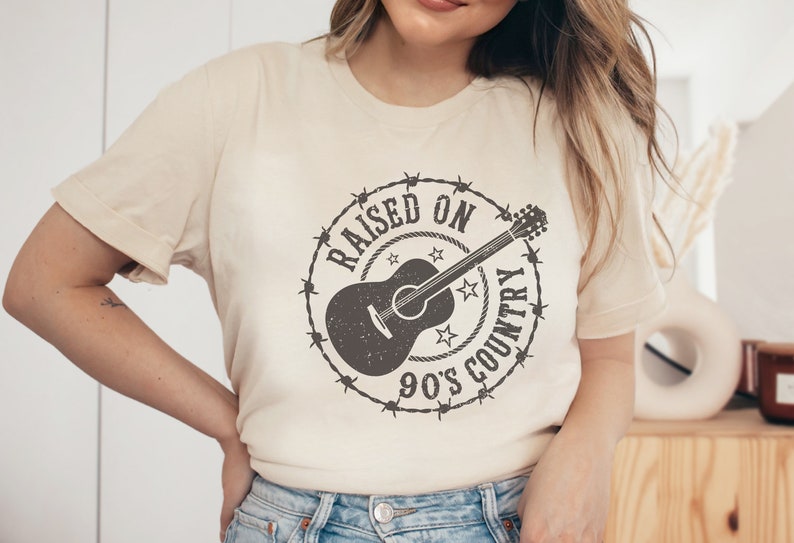 May include: Cream-colored t-shirt featuring a distressed graphic. The design includes a guitar encircled by barbed wire, stars, and the text "RAISED ON 90'S COUNTRY". A casual, short-sleeved tee.