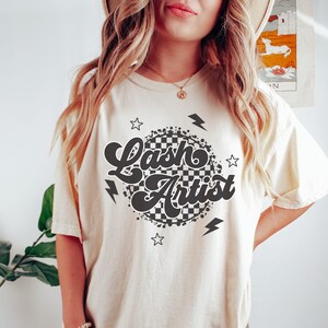 May include: Cream-colored t-shirt with the words "Lash Artist" in a retro font, surrounded by a checkered circle, stars, and lightning bolts. The shirt is a relaxed fit.