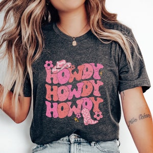 May include: A dark grey t-shirt featuring a pink and white graphic design that says "Howdy Howdy Howdy" with a pink cowboy hat and boot.
