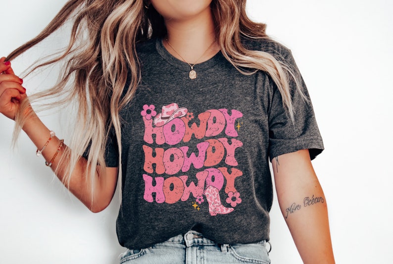 May include: A dark grey t-shirt featuring a pink and white graphic design that says "Howdy Howdy Howdy" with a pink cowboy hat and boot.