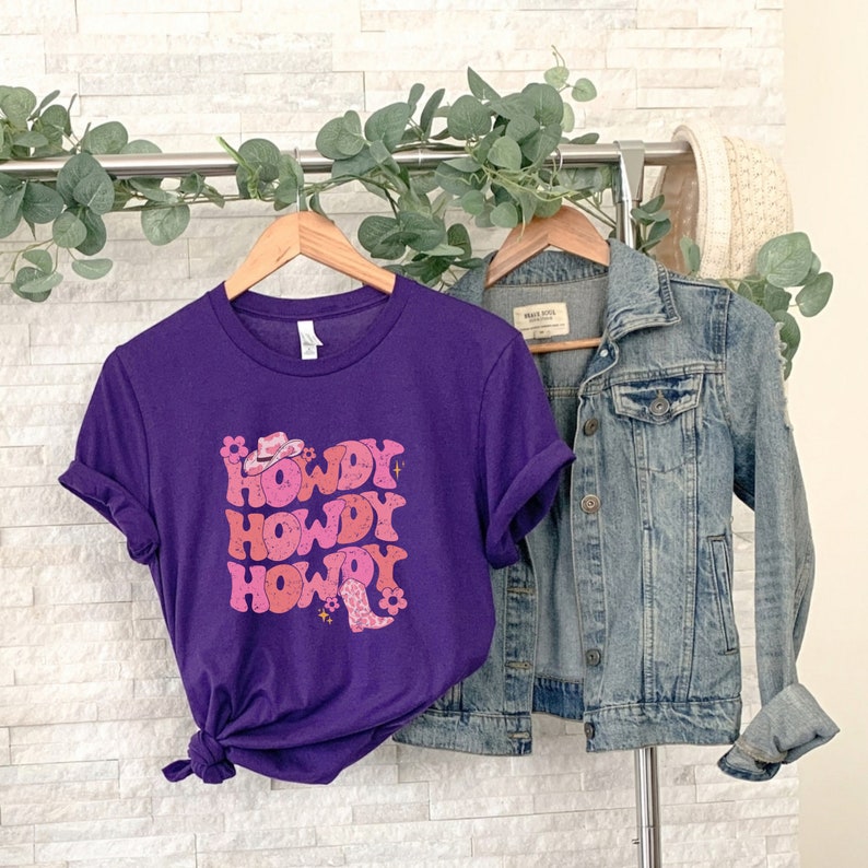 May include: Purple short-sleeved t-shirt with a pink graphic that says "Howdy" three times. The graphic also includes a pink cowboy hat and boot.