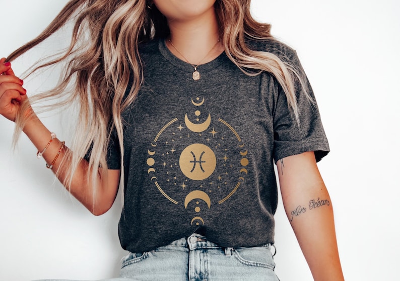 May include: A dark grey t-shirt with a gold graphic of the Pisces zodiac sign surrounded by moons and stars. The text "Pisces" is written on the shirt.
