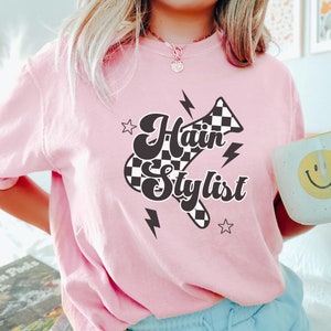 May include: Pink t-shirt with a black and white checkered hairdryer graphic and the text "Hair Stylist".