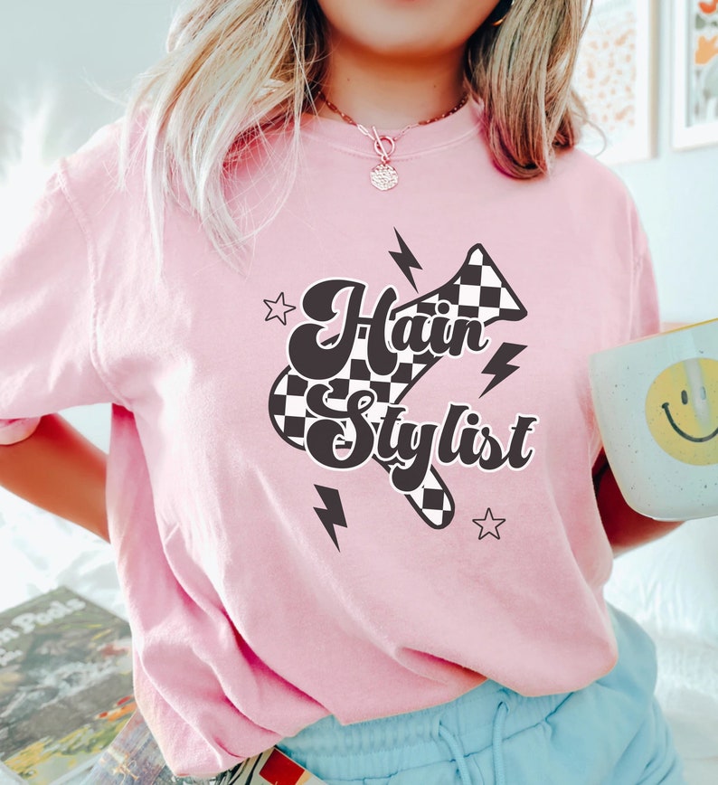 May include: Pink t-shirt with a black and white checkered hairdryer graphic and the text "Hair Stylist".