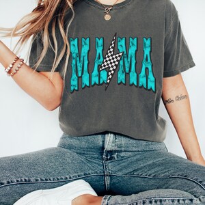 Puede incluir: Una camiseta gris con un gr&aacute;fico azul turquesa que dice "MAMA" con un rayo a cuadros en el medio.