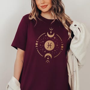 May include: A maroon t-shirt with a gold zodiac symbol for Pisces surrounded by moons and stars.