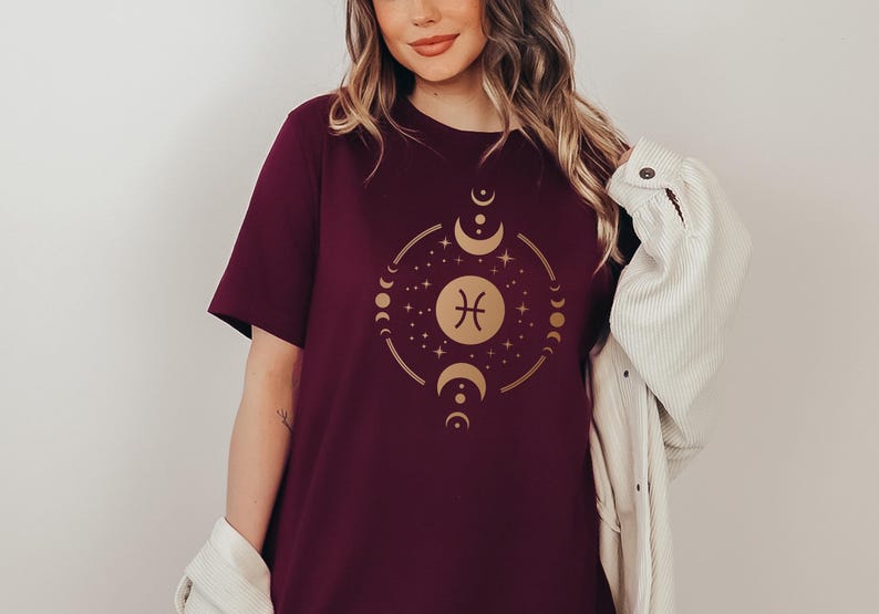 May include: A maroon t-shirt with a gold zodiac symbol for Pisces surrounded by moons and stars.