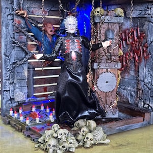 Könnte beinhalten: Eine Pinhead-Actionfigur in einem Diorama mit einer steinartigen Wand, auf der die Schriftzüge "HELL" und "TIME" zu sehen sind, hängenden Ketten, einem Haufen Totenköpfe, kleinen rotspitzigen Kerzen und einer blau beleuchteten Szenerie.