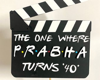 May include: A black and white clapperboard with the text "THE ONE WHERE PRABHA TURNS '40'". The clapperboard has a black base with white lettering and a black and white striped top section. The letters of the name "PRABHA" are in the colors yellow, blue, red, and blue.