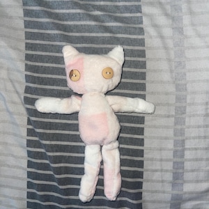 May include: A handmade plush cat doll with a white body and pink accents. The cat has button eyes and outstretched arms and legs. The doll is made of soft fabric and is designed to be cuddly.