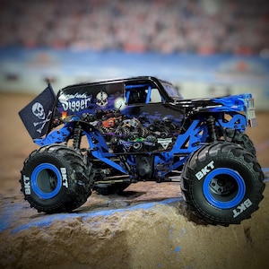 Puede incluir: Un camión monstruo negro y azul con una bandera de calavera y huesos cruzados, las palabras "Grave Digger" y "Monster Jam" en el lateral, y "BKT" en las llantas.