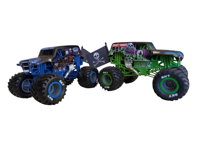 May include: Two toy monster trucks, one blue and one green, are displayed. The blue truck has a pirate flag and the text "Grave Digger" on the green truck. Both have large tires with the text "BKT".