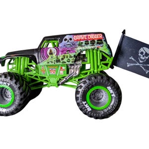 May include: A Grave Digger monster truck toy with a black and green color scheme. The truck features large tires with BKT branding, a skull graphic, and a pirate flag. The text "GRAVE DIGGER" is visible on the roof.