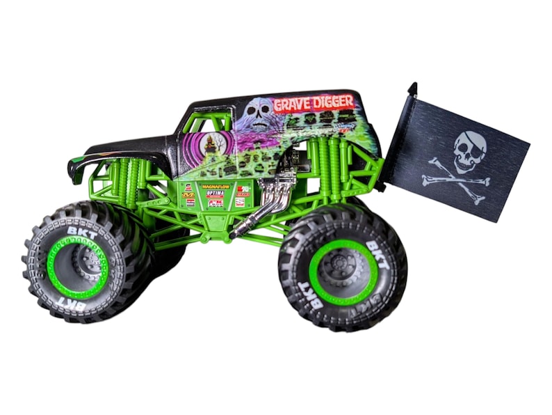 May include: A Grave Digger monster truck toy with a black and green color scheme. The truck features large tires with BKT branding, a skull graphic, and a pirate flag. The text "GRAVE DIGGER" is visible on the roof.