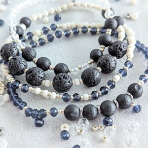 Peut inclure: Un ensemble de trois bracelets avec des perles de lave noire, des perles blanches et des perles de verre bleu foncé. Les bracelets sont superposés sur une surface blanche.