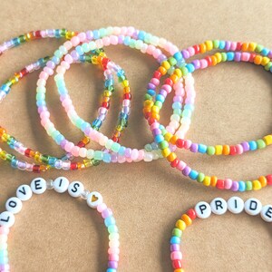 May include: Five rainbow beaded bracelets with a small heart charm and two bracelets with the words "LOVE IS" and "PRIDE" spelled out in white letters.