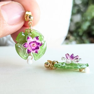 Peut inclure: Une paire de boucles d'oreilles en forme de feuille verte avec une fleur rose et violette. Les boucles d'oreilles sont en résine et ont une finition dorée.