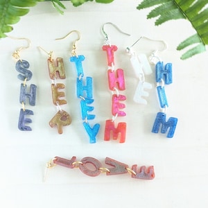 May include: A set of dangle earrings with the words "SHE", "THEY", "HIM", and "THEM" spelled out in colourful letters. There is also a separate earring with the word "LOVE" spelled out in colourful letters.
