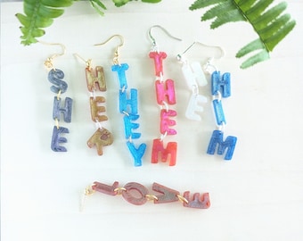 May include: A set of dangle earrings with the words "SHE", "THEY", "HIM", and "THEM" spelled out in colorful letters. There is also a separate earring with the word "LOVE" spelled out in colorful letters.