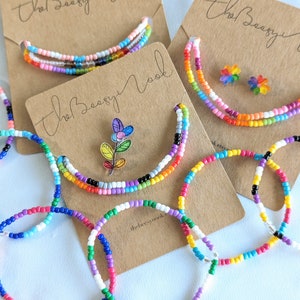 May include: A set of colourful beaded bracelets and necklaces with a flower charm. The bracelets and necklaces are made with small, colourful beads and are arranged on a brown background with the text "The Beazy Nook".