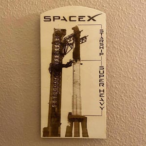 May include: A wooden sign with a black and white image of a SpaceX Starship and Super Heavy rocket on a launchpad. The sign reads "SPACEX" at the top and "STARSHIP - SUPER HEAVY" on the right side.