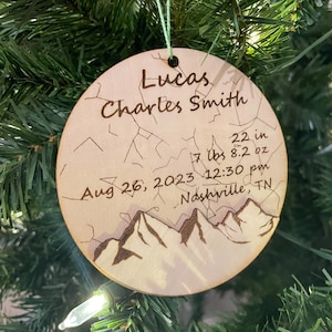 May include: A round wooden ornament with a light brown background and a mountain range design. The ornament is personalized with the name "Lucas Charles Smith", birth date "Aug 26, 2023", time "12:30 pm", location "Nashville, TN", weight "7 lbs", and height "22 in".