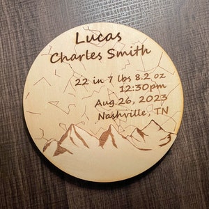 May include: A round wooden plaque with a mountain range design and the text "Lucas Charles Smith", "22 in 7 lbs 8.2 oz", "12:30pm", "Aug 26, 2023", and "Nashville, TN".