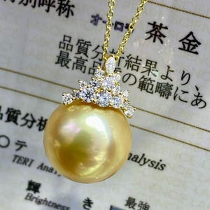 May include: A gold necklace featuring a large, lustrous golden pearl pendant. The pendant is adorned with a cluster of small, sparkling diamonds. The chain is delicate and gold. The background includes Japanese text and a grading report.