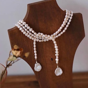 Op de afbeelding: Een parelketting met drie strengen, tentoongesteld op een houten sieradenstandaard. De ketting heeft kleine, crèmekleurige parels en twee grote, onregelmatig gevormde parelhangers. De houten standaard is donkerbruin.