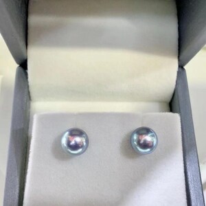 Puede incluir: Un par de pendientes de perlas redondas e iridiscentes presentados en una caja de joyer&iacute;a gris con interior blanco. Las perlas tienen un sutil tono azul-gris y una superficie reflectante, lo que sugiere alta calidad.