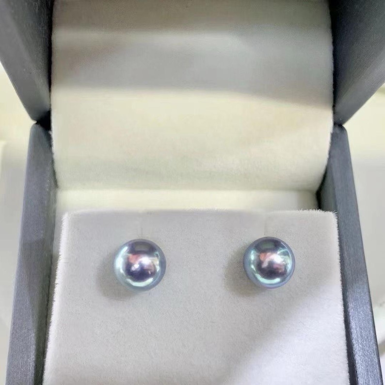 Puede incluir: Un par de pendientes de perlas redondas e iridiscentes presentados en una caja de joyer&iacute;a gris con interior blanco. Las perlas tienen un sutil tono azul-gris y una superficie reflectante, lo que sugiere alta calidad.
