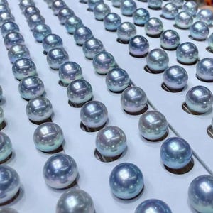 Puede incluir: Filas de perlas redondas e iridiscentes en tonos azules, plateados y blancos, dispuestas sobre una superficie blanca. Las perlas est&aacute;n espaciadas uniformemente y tienen una superficie lisa y reflectante.