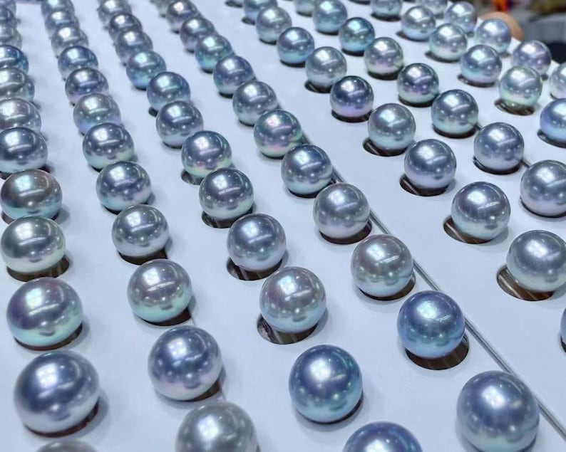 Puede incluir: Filas de perlas redondas e iridiscentes en tonos azules, plateados y blancos, dispuestas sobre una superficie blanca. Las perlas est&aacute;n espaciadas uniformemente y tienen una superficie lisa y reflectante.