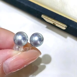 Puede incluir: Un par de pendientes de perlas grises. Las perlas son redondas y tienen un brillo lustroso. Los pendientes est&aacute;n engarzados en postes plateados con cierres transparentes. El fondo incluye una caja de joyer&iacute;a azul oscuro y un cierre dorado.