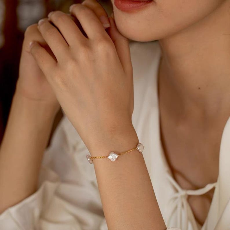 K&ouml;nnte beinhalten: Ein goldfarbenes Armband mit vier wei&szlig;en Kleeblatt-Anh&auml;ngern und drei unregelm&auml;&szlig;ig geformten Perlen. Das Armband wird am Handgelenk getragen, wobei die H&auml;nde und ein Teil des Gesichts der Person sichtbar sind.