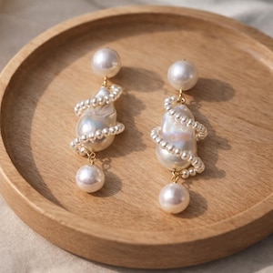 May include: A pair of elegant pearl earrings. Each earring features a large, irregular-shaped pearl wrapped with a strand of smaller pearls. A smaller pearl dangles below, and a round pearl tops each earring. The earrings are gold-toned.