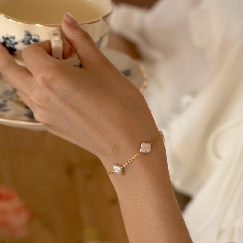 K&ouml;nnte beinhalten: Ein zartes goldenes Armband mit vier unregelm&auml;&szlig;ig geformten, wei&szlig;en S&uuml;&szlig;wasserperlen. Das Armband ist durch eine feine Goldkette verbunden. Der Hintergrund zeigt eine Tasse und Untertasse mit floralem Design.